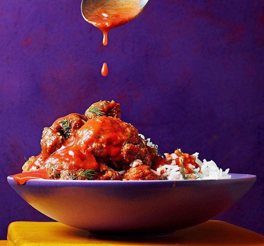 Red sauce dripping onto cooked meatballs with rice in bowl against purple background