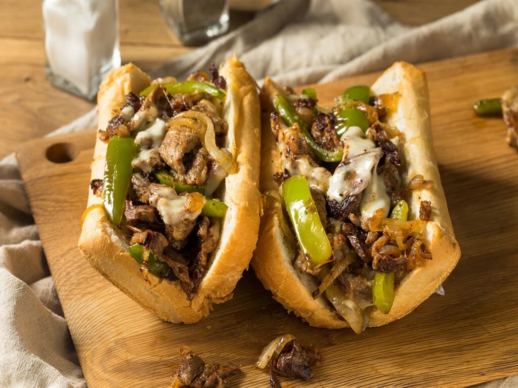 Two Philly cheesesteak sandwiches with sliced beef, melted cheese, and green peppers on wooden board