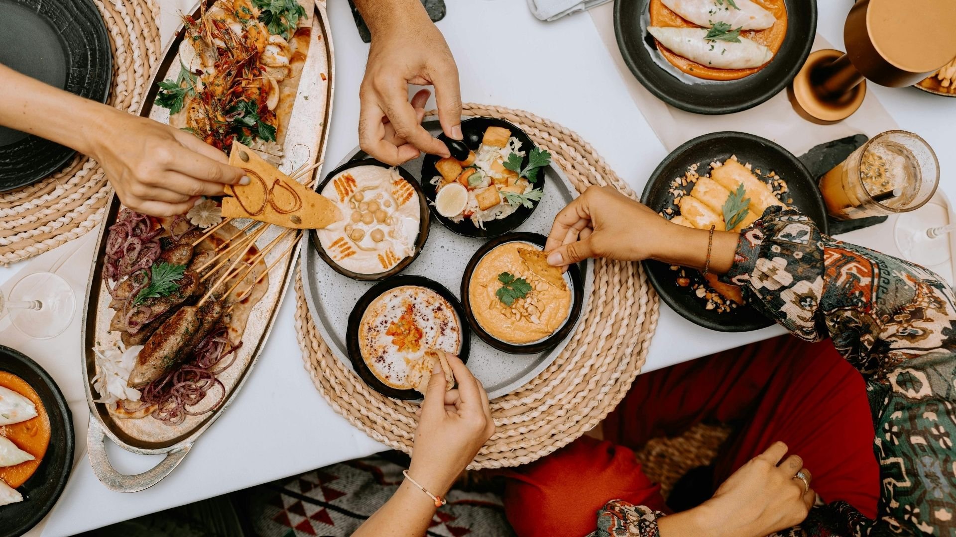 Overhead view of people sharing Mediterranean-style meal with hummus, meats, and fresh herbs.
